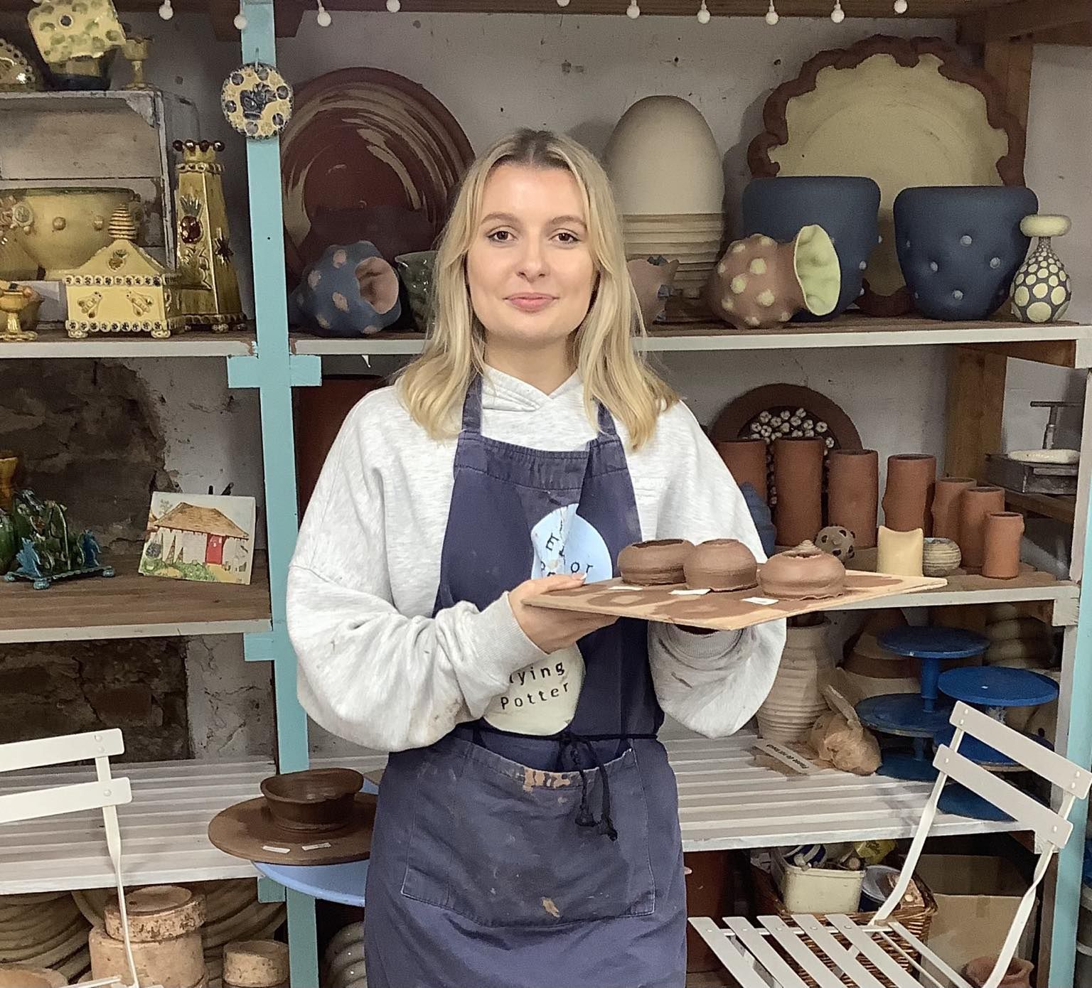 young woman holding up a board of freshly thrown pots made on the potter's wheel. she is looking at the camera and smiling