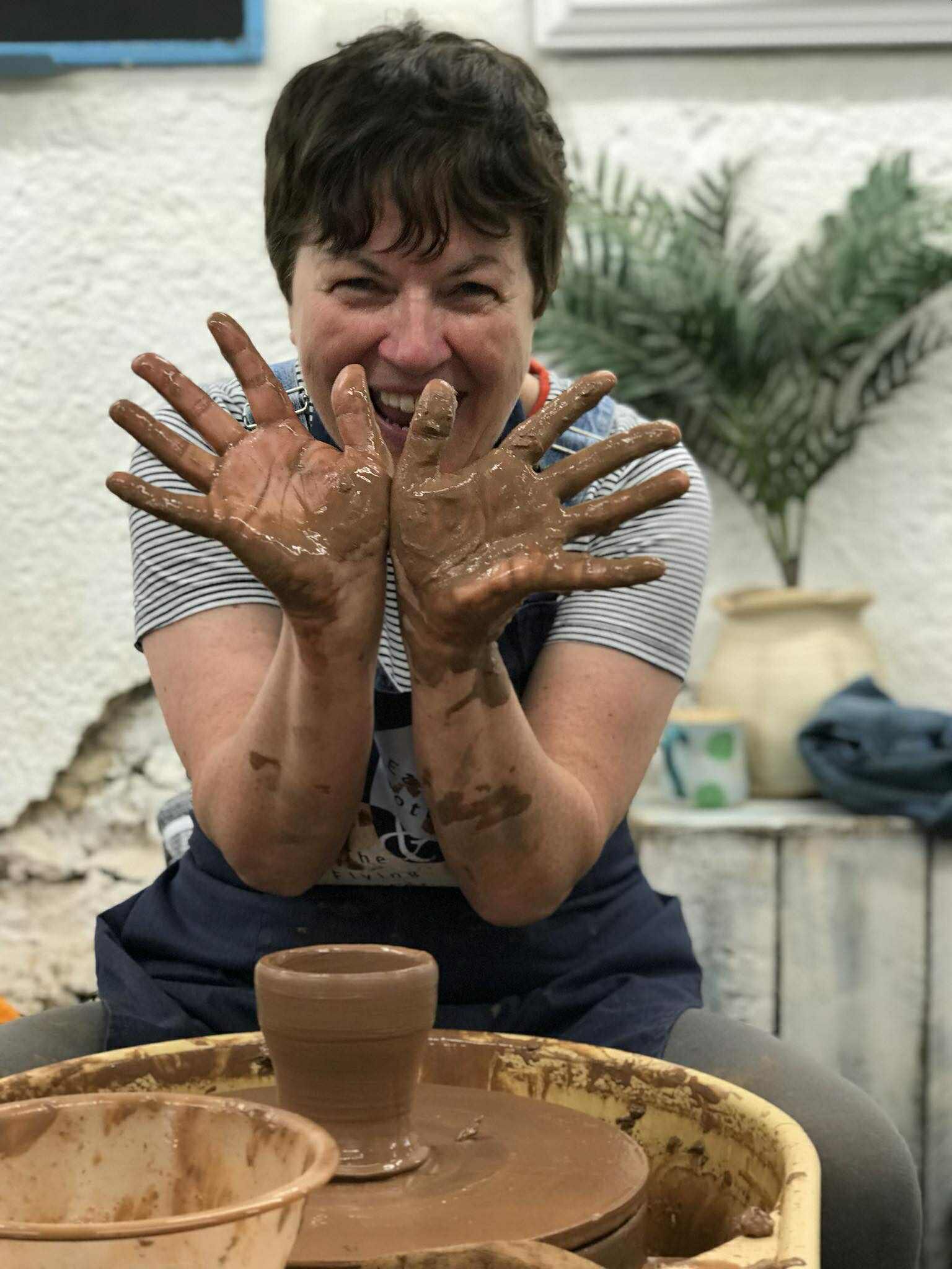 A lady having a wonderful time sat at a pottery wheel. She has a freshly made pot in the centre of her wheel in the shape of a beaker. she is holding her hands up in front of her face showing her hands covered in clay.