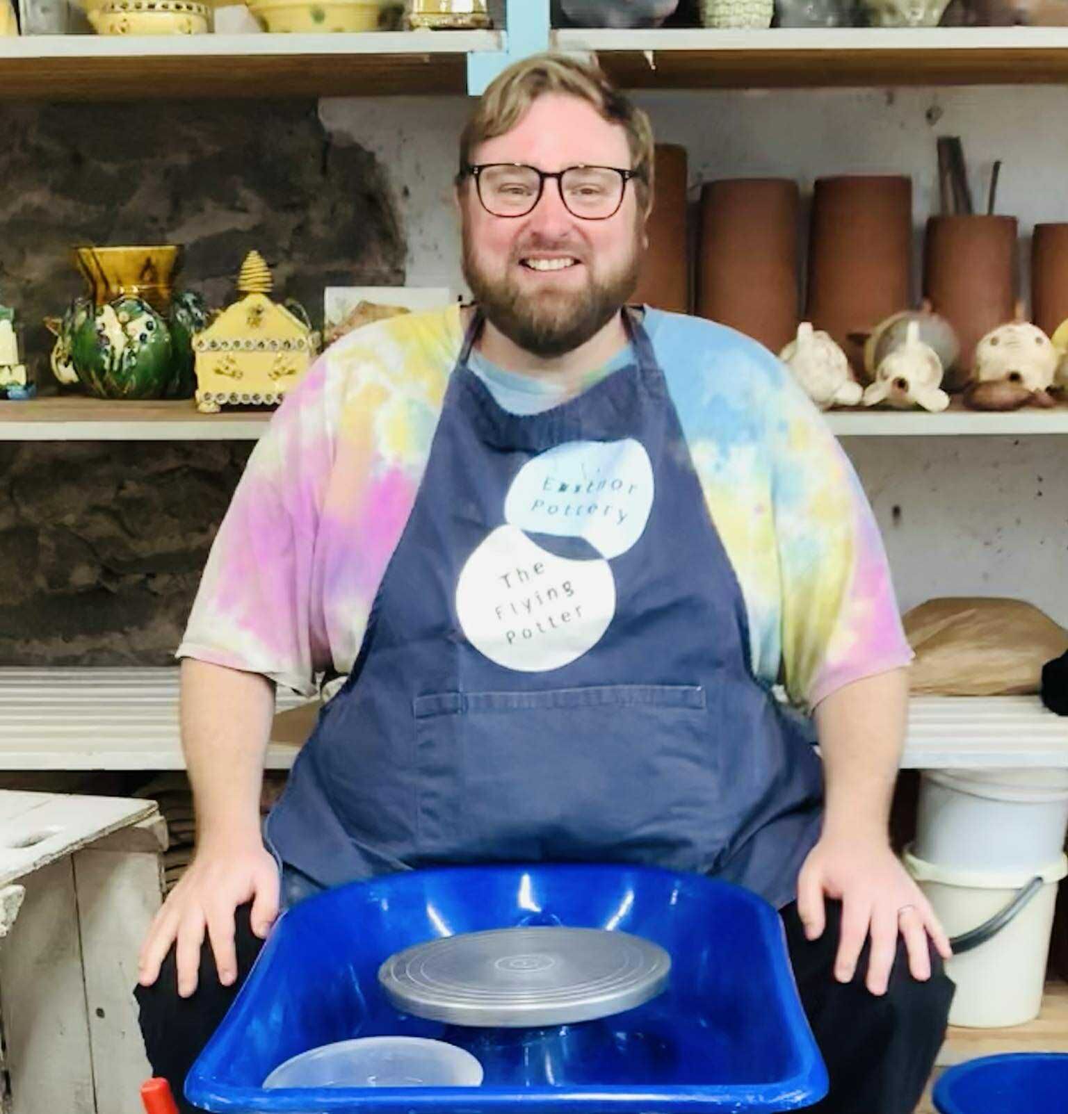 Male potter's wheel course participant wearing a pale tied dyed t-shirt and navy blue eastnor pottery apron with logo. He's sat smiling with hands on legs on a clean potter's wheel before the class began.