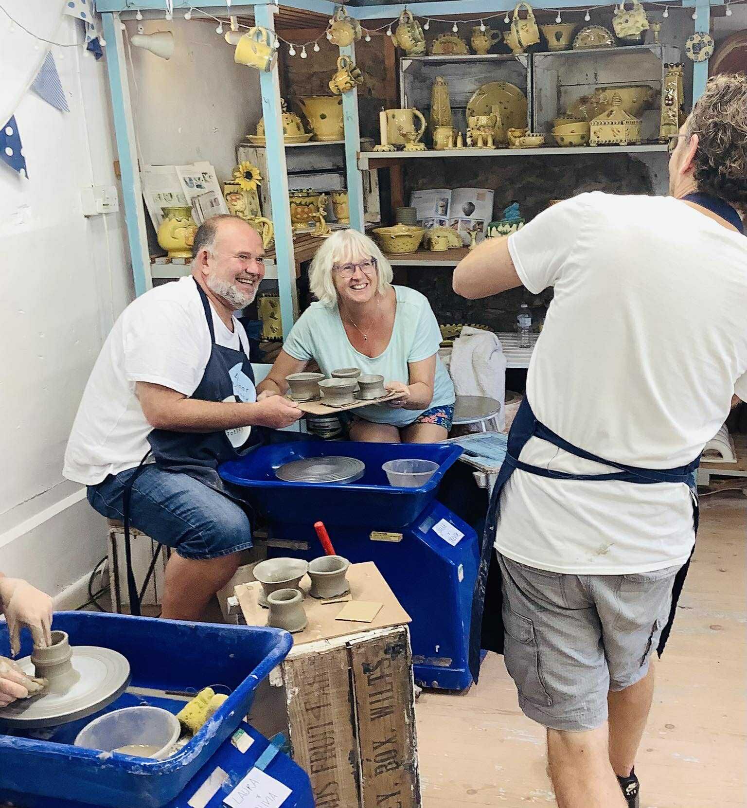 A grey haired couple sitting at a pottery wheel, they are holding up a board of 4 grey freshly made pots they are smiling at a man who is taking a photograph with his back to the camera. The couple are smiling at the camera.