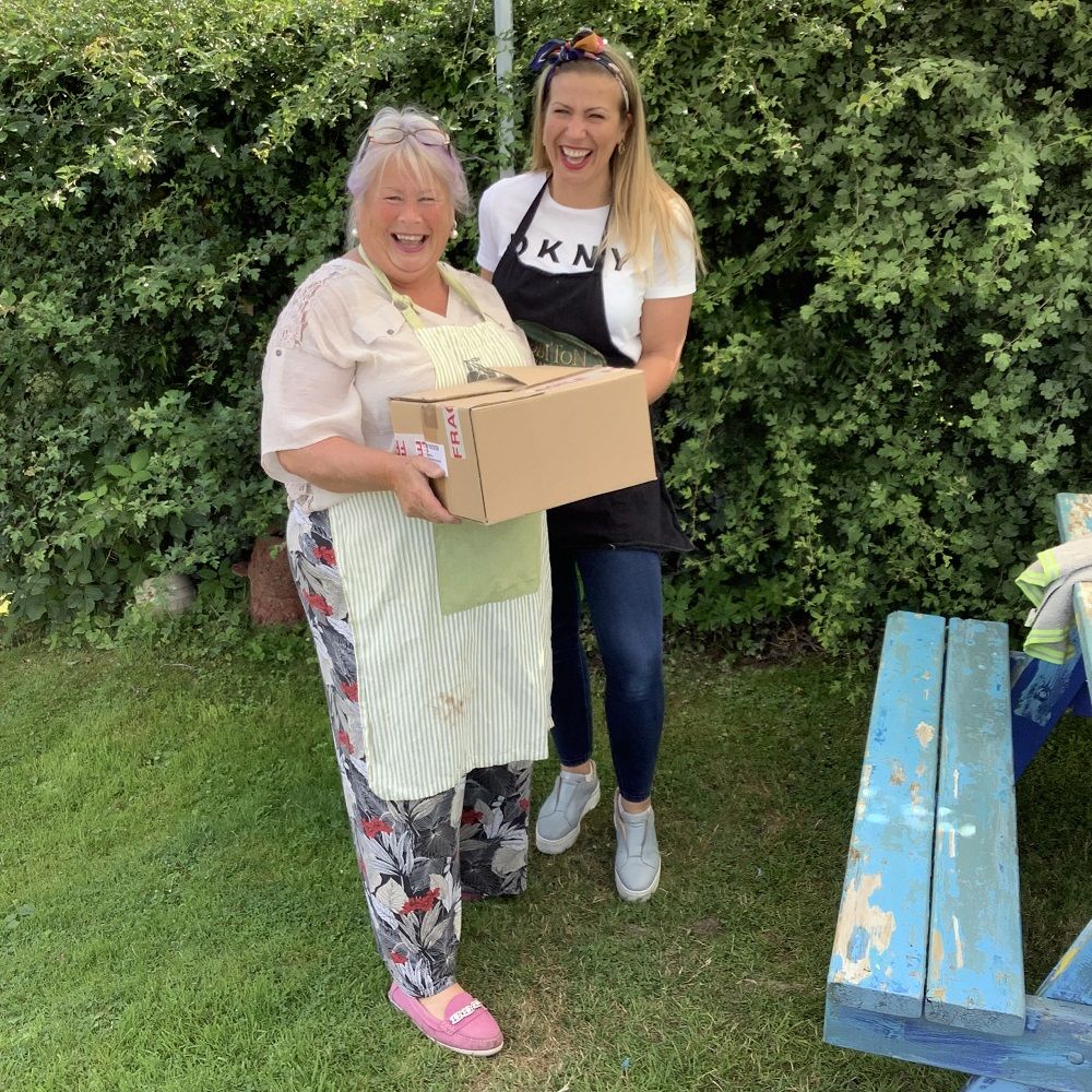 mother and daughter holding cardboard box smiling, in front of hedgerow 