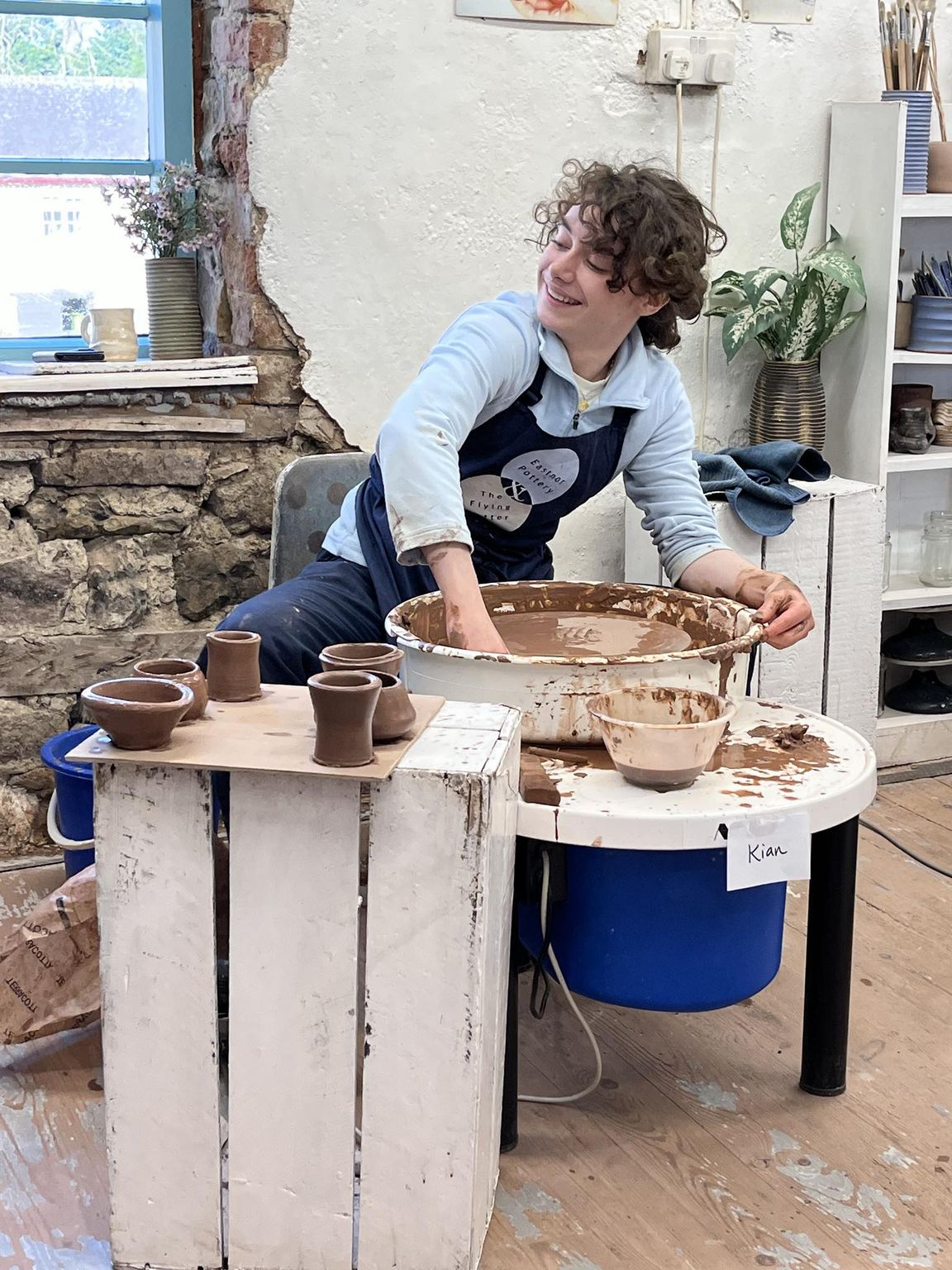 teenager sat smiling and looking to his right as he cleans his potter's wheel. There is a board of freshly thrown, mug sized pots on a board to the front of the composition