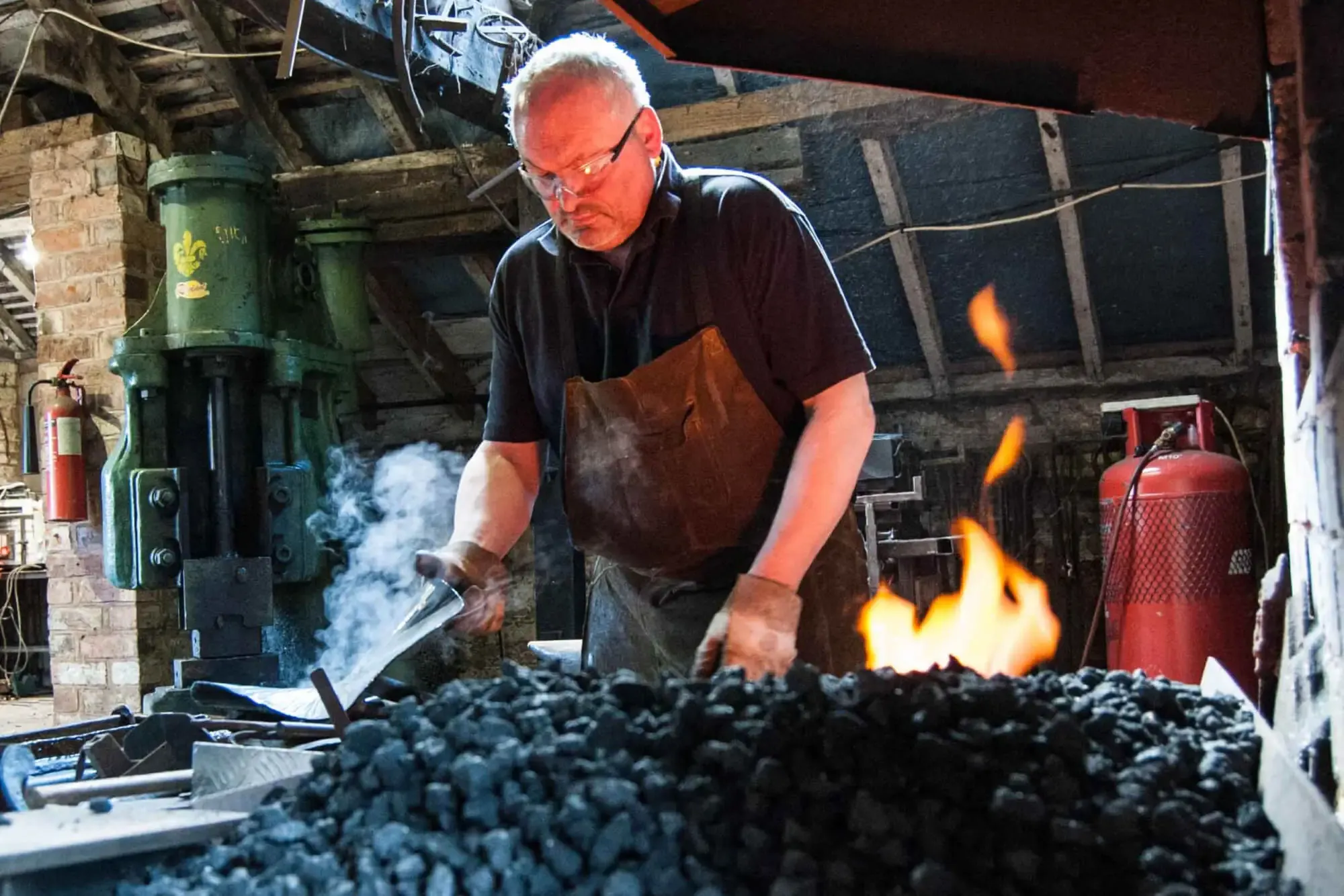 Andrew Findlay black smith at work, leaning over hot coals, there are flames. He is holding a piece of steaming metal. He is wearing protective goggles, leather gloves and apron. In the background is a large red gas cannister, blacksmith equipment and a fire extinguisher.