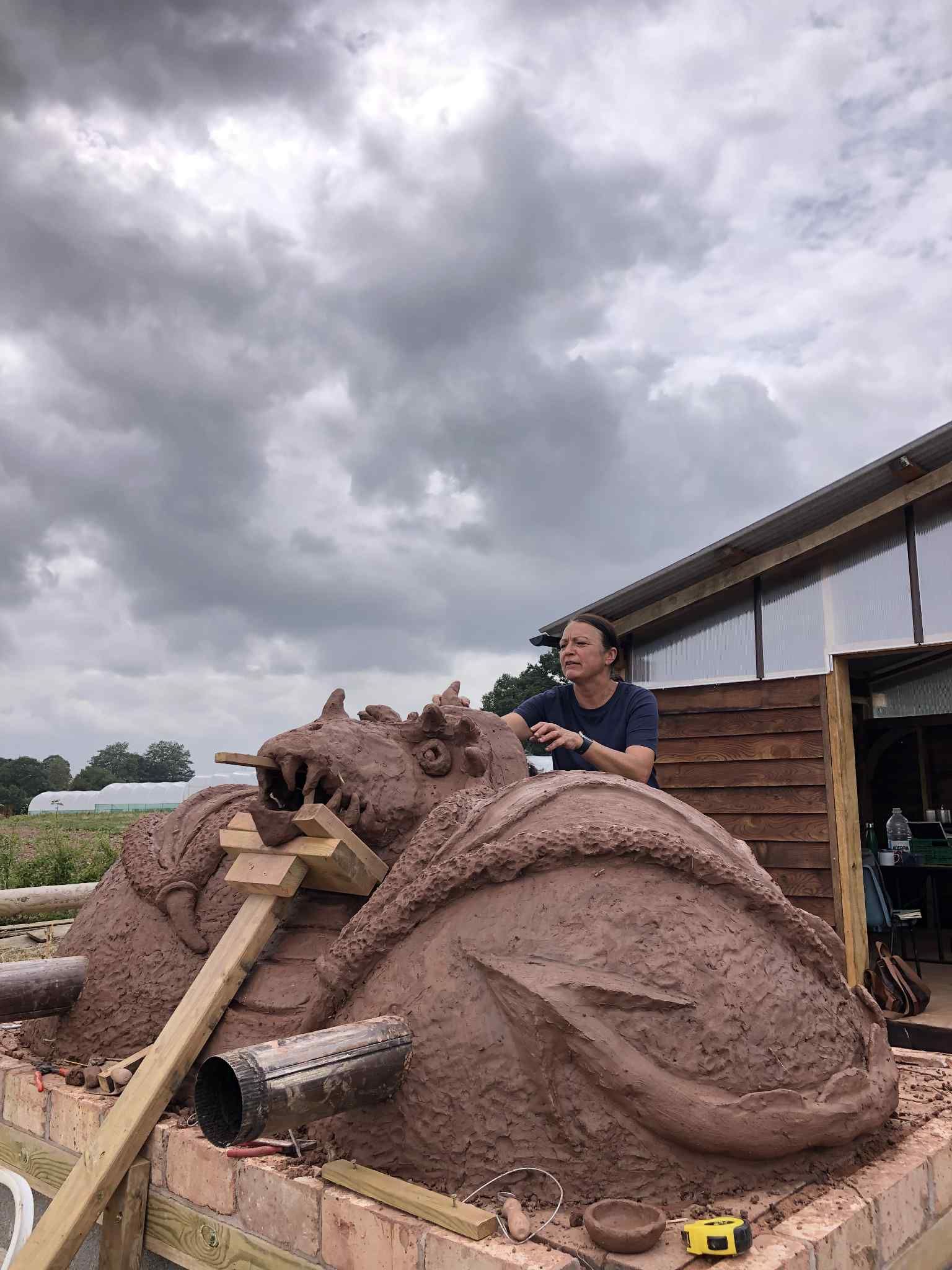 front of cob pizza oven in the shape of a dragon with it's mouth open. Dragon chin is supported by a wooden brace to prevent it from collapsing