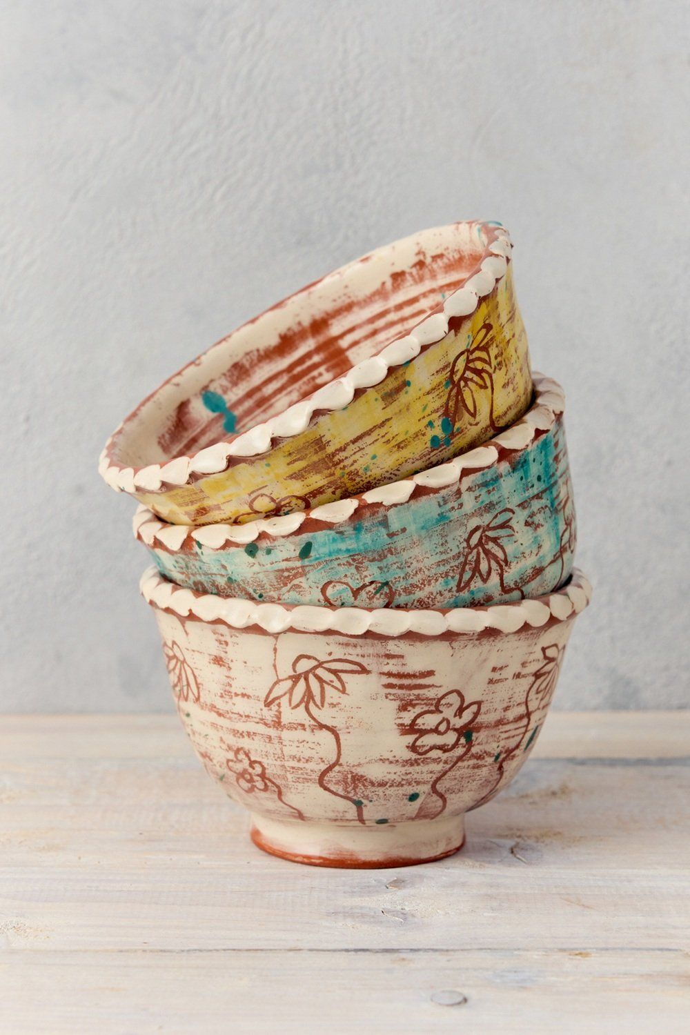 Three stacking bowls made by Sarah Monk. Each bowl is the same size, but stacked up inside each other. They are decorated individually with sgraffito flowers. A white bowl at the bottom, a teal bowl in the centre and a yellow bowl at the top. The pots have white pie-crust type rims.