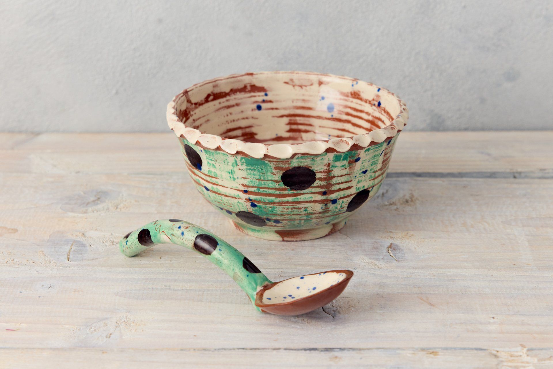 A pottery soup bowl with matching pottery spoon. The bowl is green with black polka dots, with a white pie crust rim. The spoon has a green handle with black dots. The spoon end is painted white. The bowl is made by Eastnor potter, Sarah Monk.