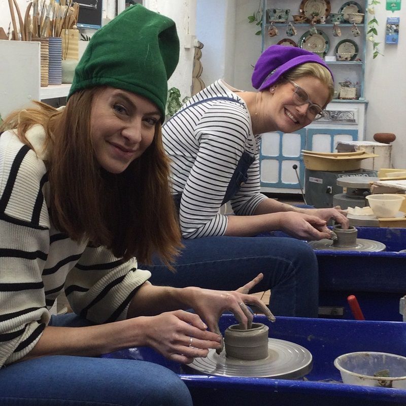 Two smiling ladies sat at the potter's wheels making a pot and wearing Christmas elf hats.