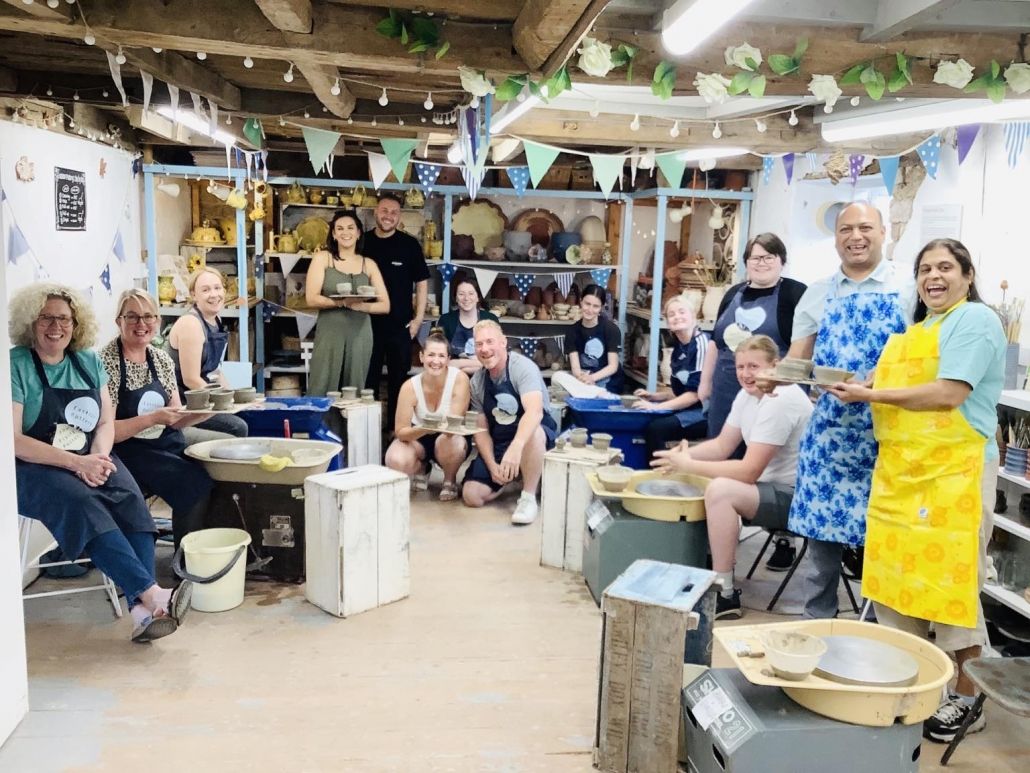 group of pottery experience participants standing by their potter's wheels proudly showing off the clay pots they have made