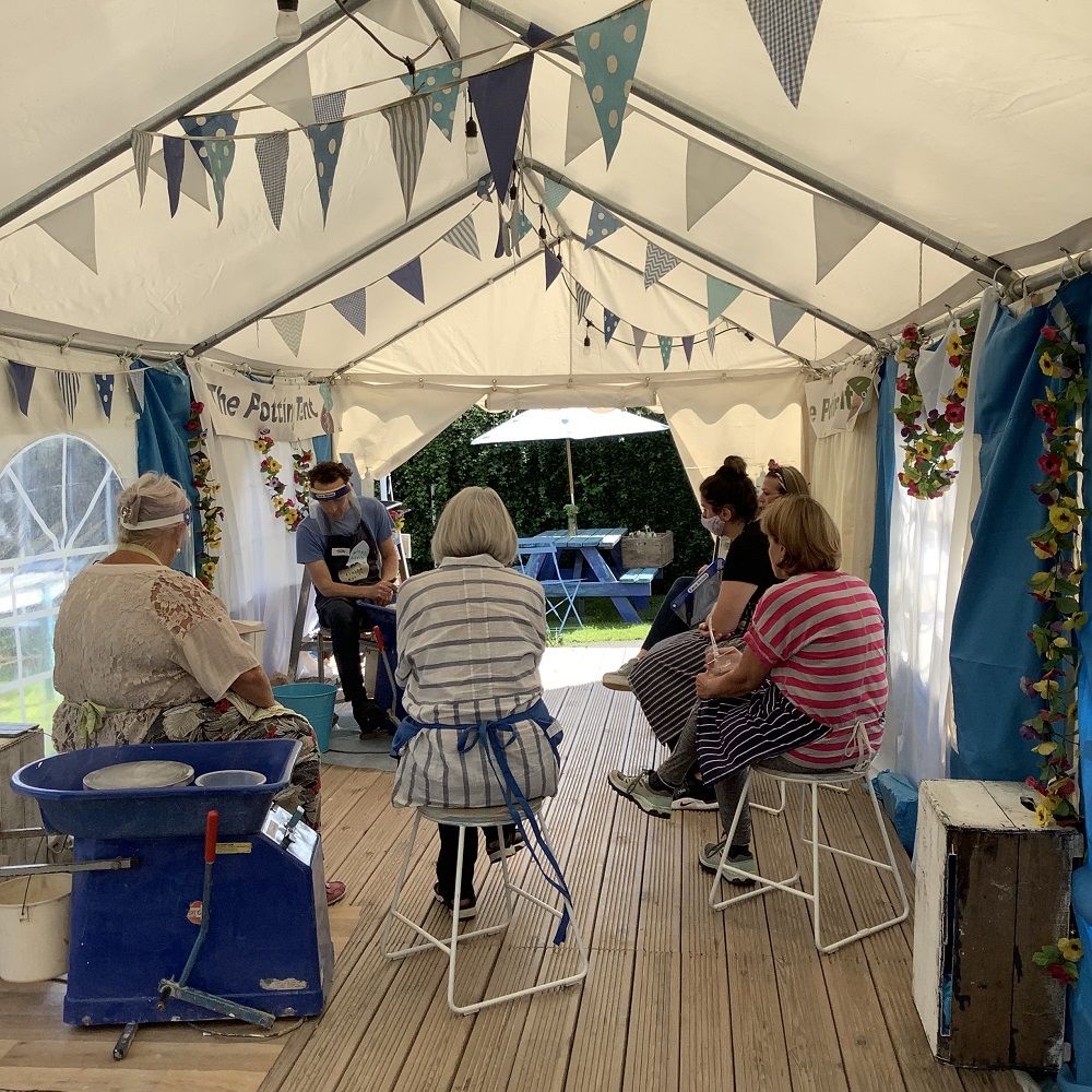 five woman sat around jon williams doing a throwing demo on a potters wheel in side a marquee