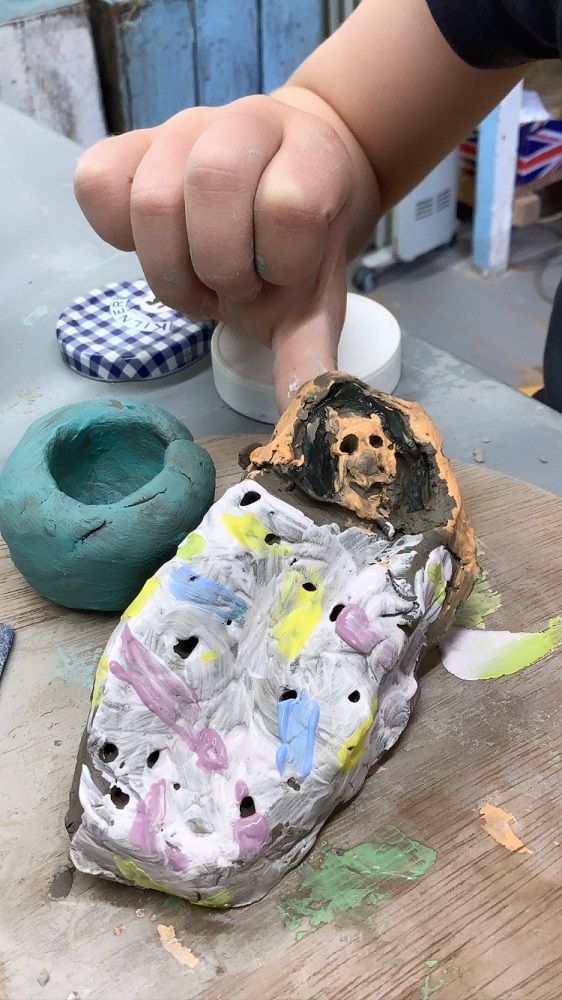 child's hand holding up clay creation of a lady inside a sleeping bag painted orange, white, pink, yellow and blue