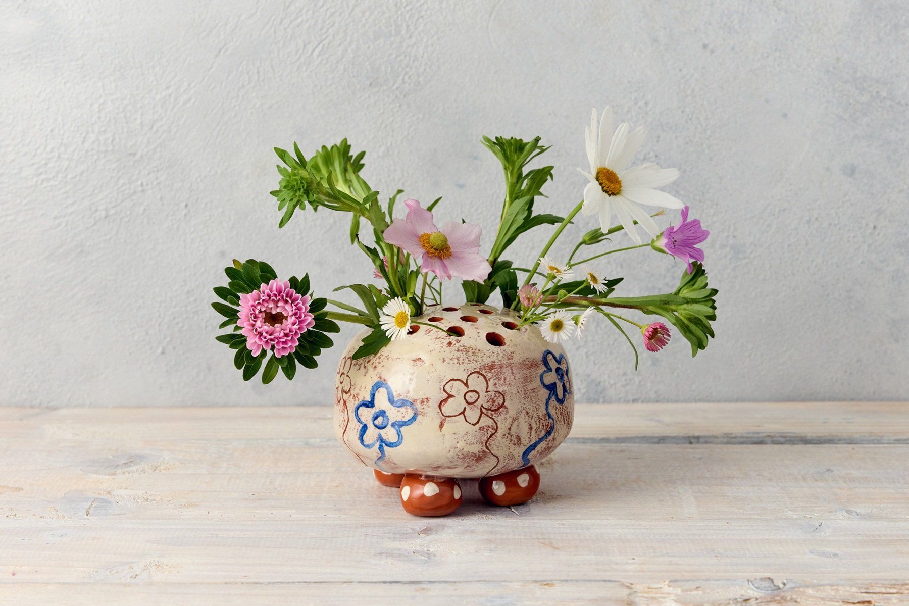 A round flower brick with holes in the top. It is white with simple painted flowers and sgraffito. The pot sits on 4 round dotty feet. The pot also contains an arrangement of fresh flowers, including daisies and cosmos.