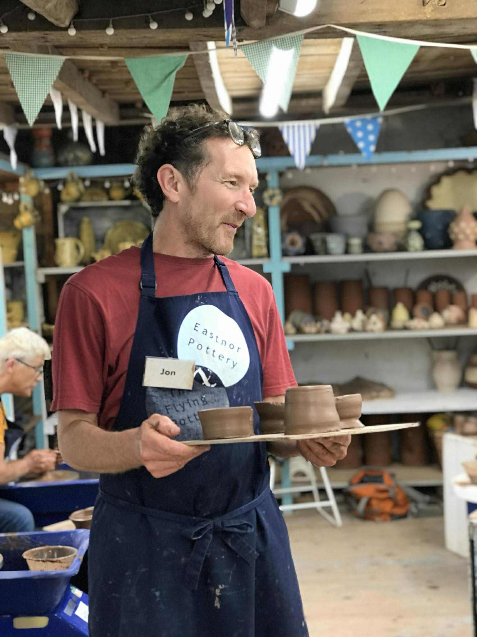 A man stood in the centre of the studio holding a board of freshly made pottery. He is wearing an Eastnor pottery apron and a name tag the say Jon.