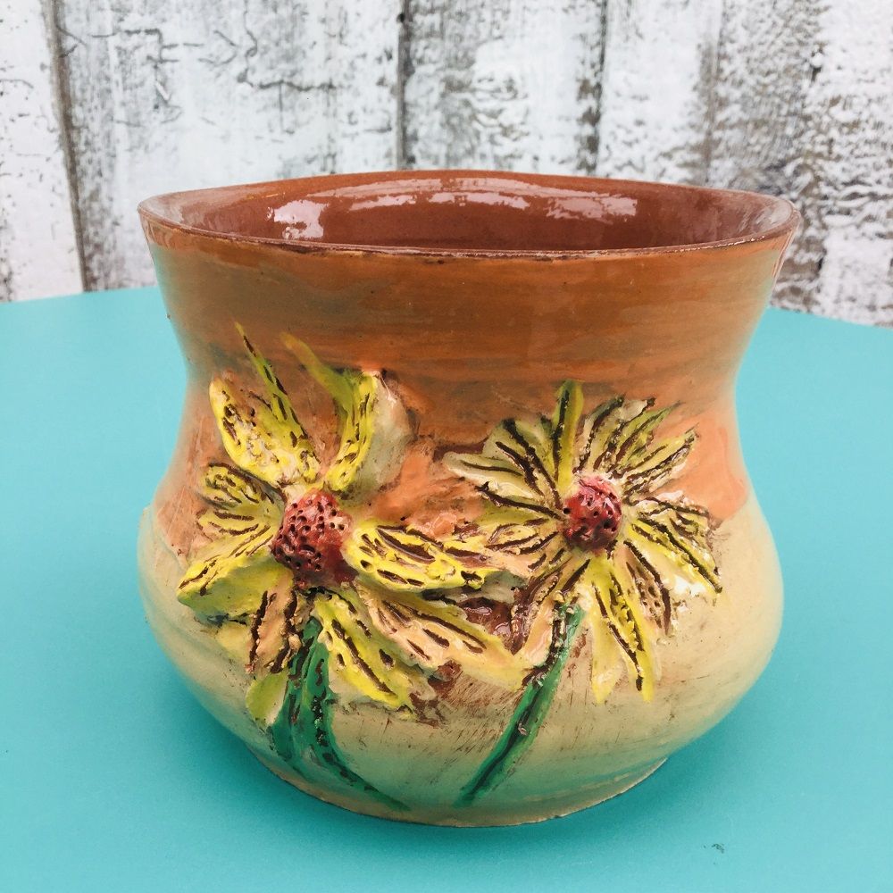 a fired and glazed hand painted terracotta vase made by a potter's wheel course participant at eastnor pottery herefordshire. There are two large modelled flowers on the side with long yellow petals and red centres. both have green stalks