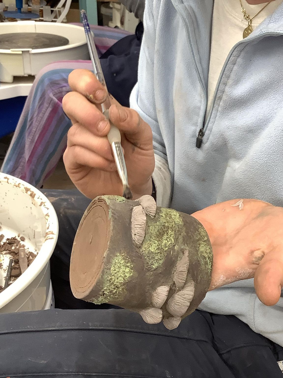 close up of a hand made mud with modelled mushrooms protruding from the surface. its being held in the left hand whilst the right hand paints detail onto the surface 