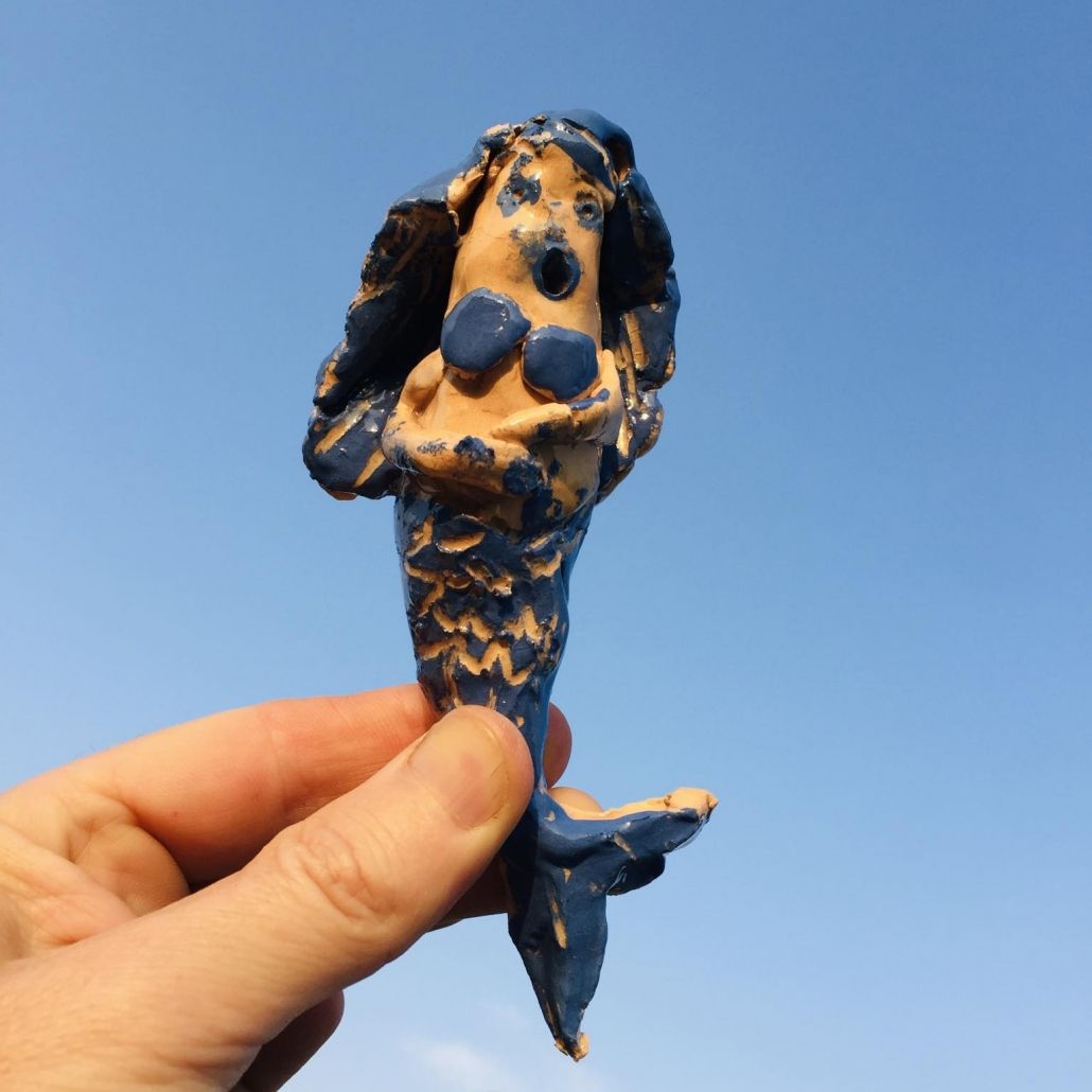 blue and tanned pottery mermaid made by a child and held by an adult hand against a blue sky background