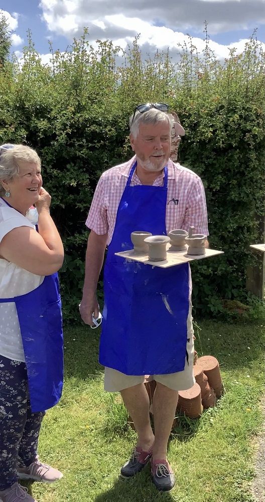 husband and wife stood outside chatting holding a wooden board with pots thrown on introduction to the potters wheel course