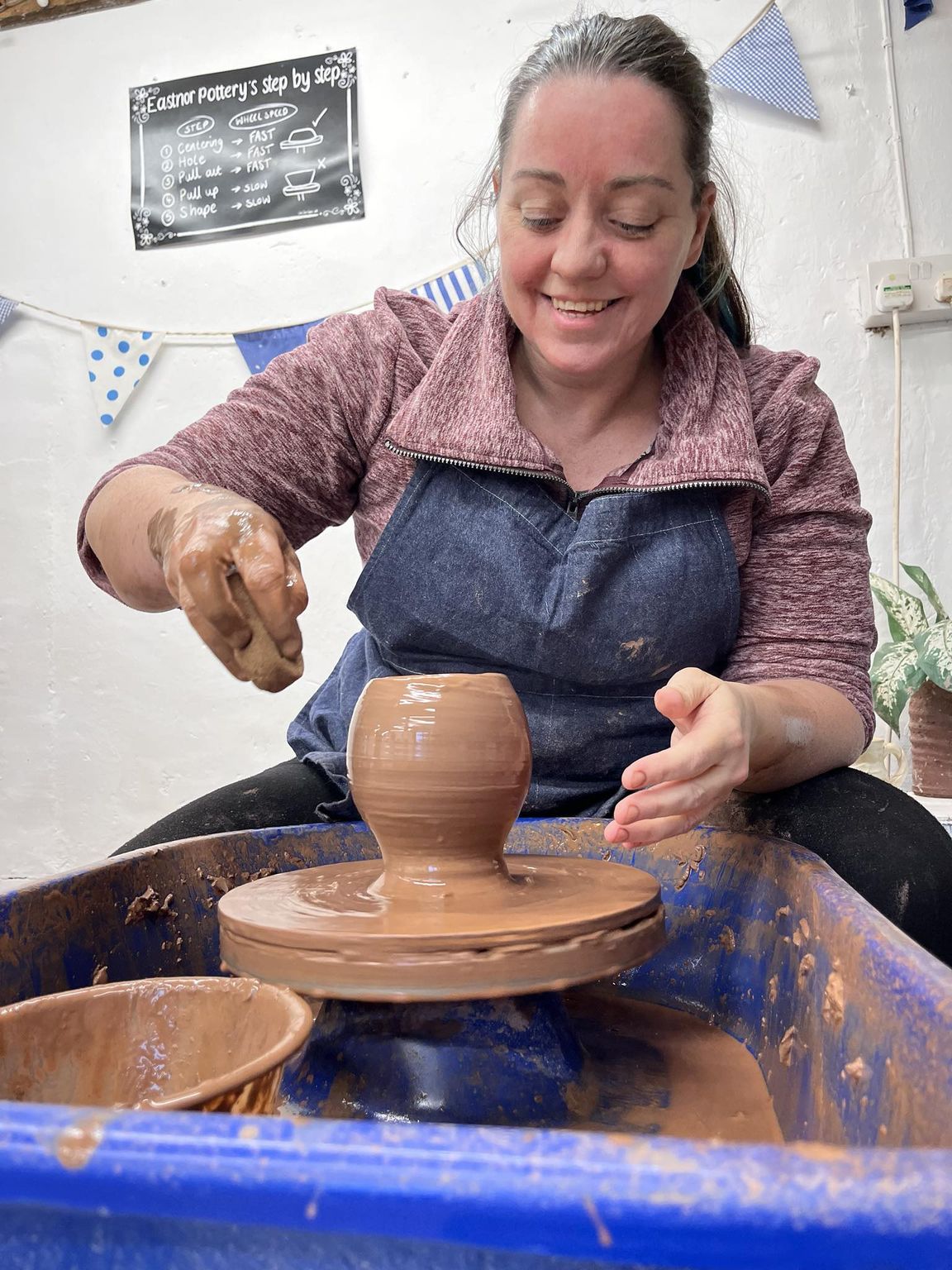 woman on a potter's wheel smiling and admiring her wet vase she'd just shaped on the potter's wheel. her right hand is holding a sponge above the pot