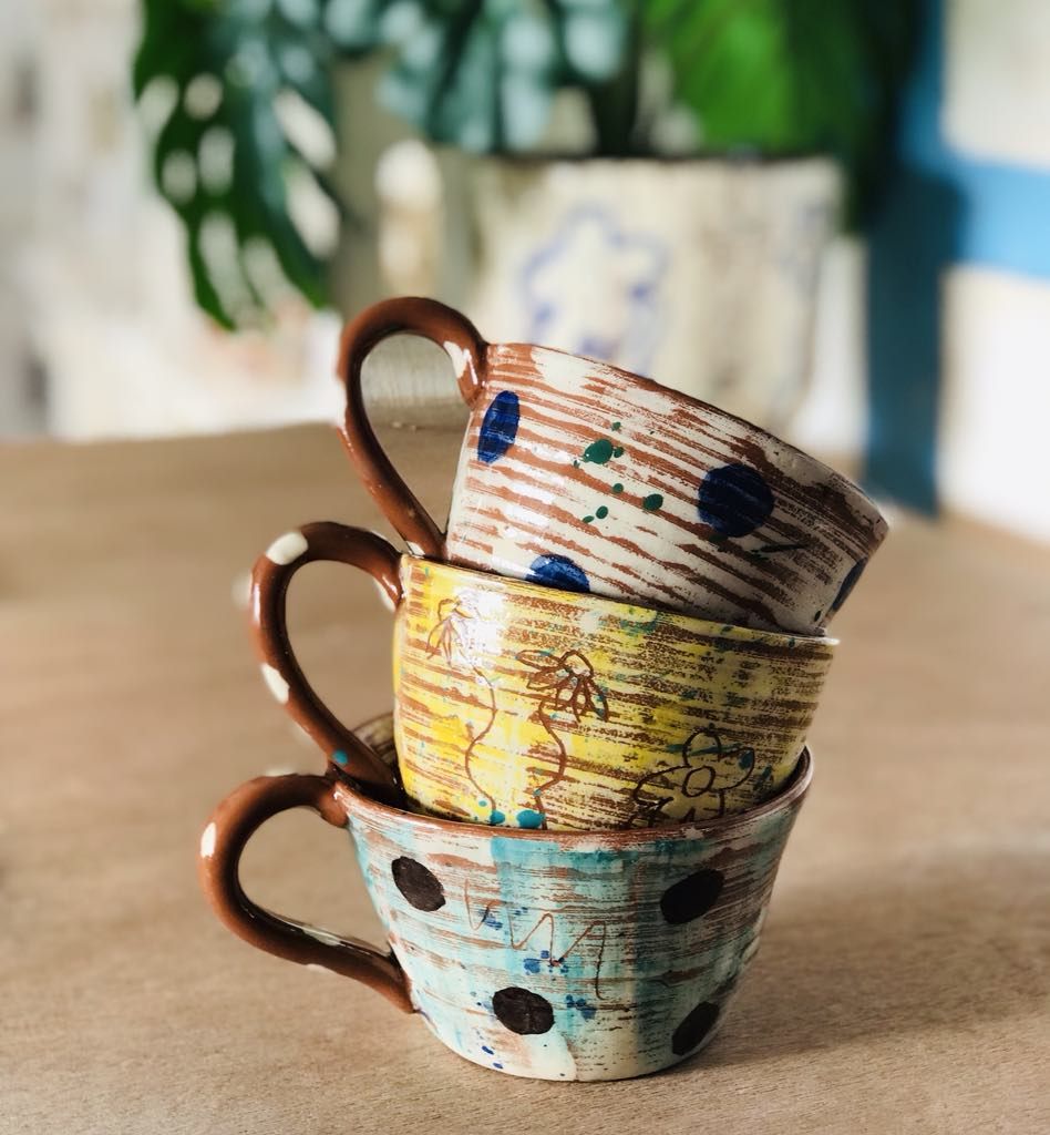 A stack of three coffee cupsthe bottom cup is teal with dark brown dots, the middle cup is yellow with sgraffito flowers and the top cup is white with cobalt blue dots. They all have terracotta handles with white painted dots. Throwing lines can be seen on all the pots. Designed and made by Sarah Monk Ceramics.