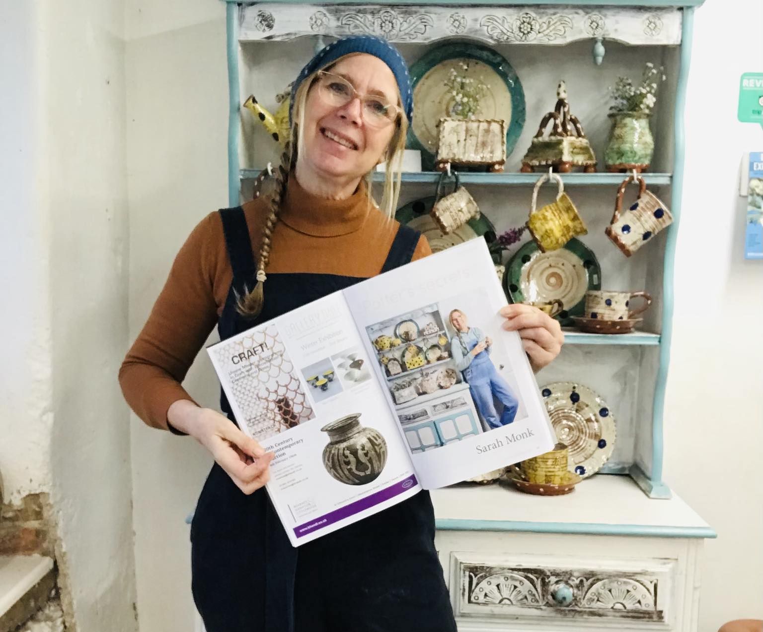 Sarah Monk Ceramics holding up an open copy of ceramic review magazine showing the article she wrote. Sarah Monk is standing in her studio at eastnor pottery in front of a dresser display of her recent pottery.