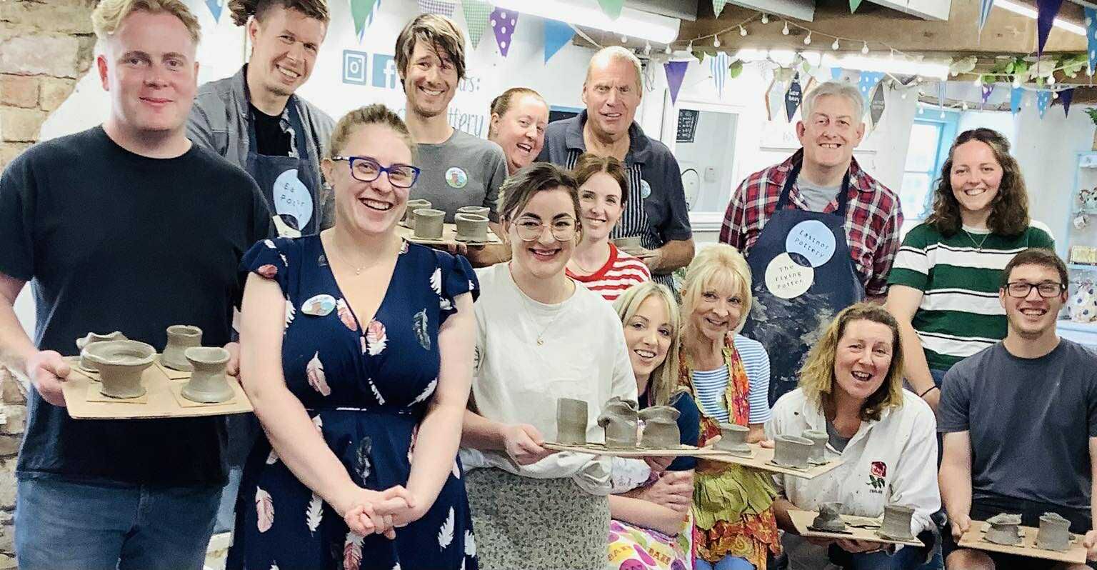 A group of happy potters, 6 men and 8 women. Some are holding boards of freshly made grey coloured pots. * are stood up whilst the rest are sat down in a row in front. Some are wearing aprons.