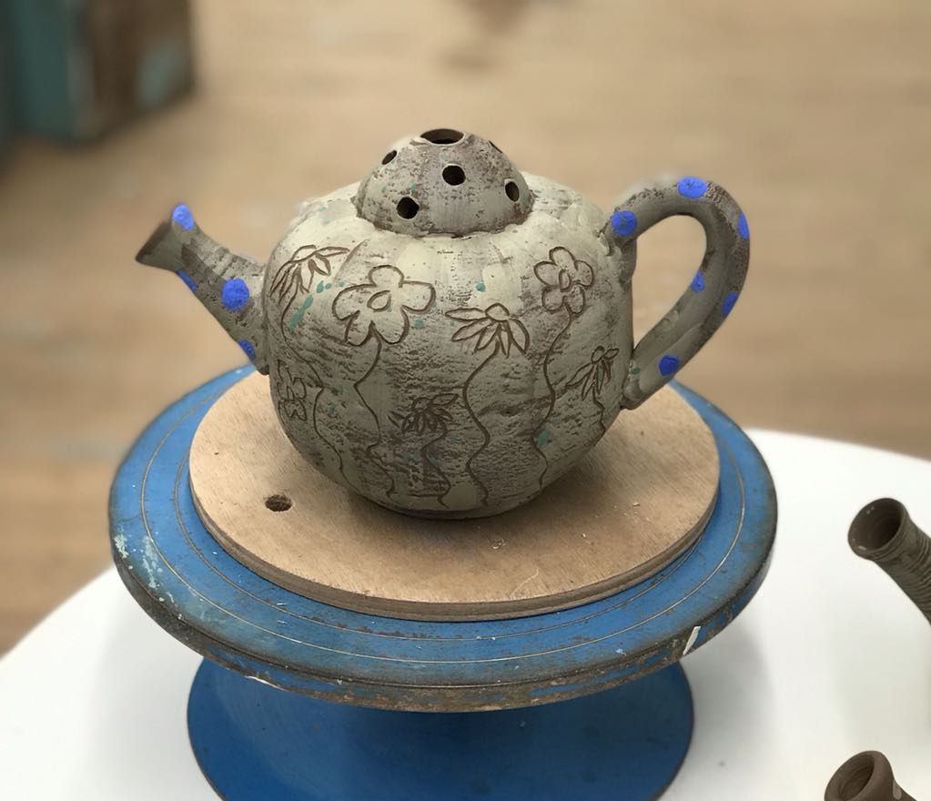 Clay teapot on the making table at Eastnor Pottery created by Sarah Monk. It is painted with white slip and decorated with sgraffito flowers. There is a lid with holes in the top to place flowers.