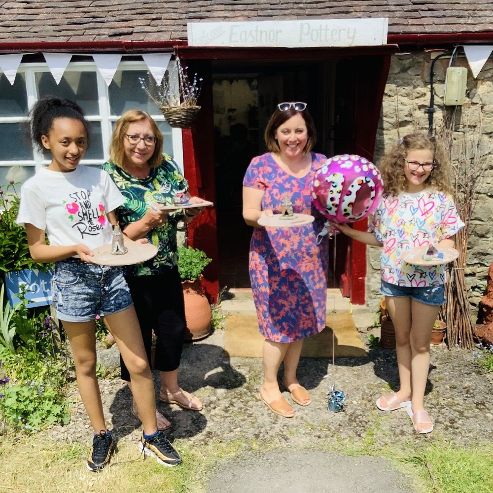 2 adults and 2 children stood outside pottery holiding 10th birthday baloon and trays with their clay creations made at eastnor pottery