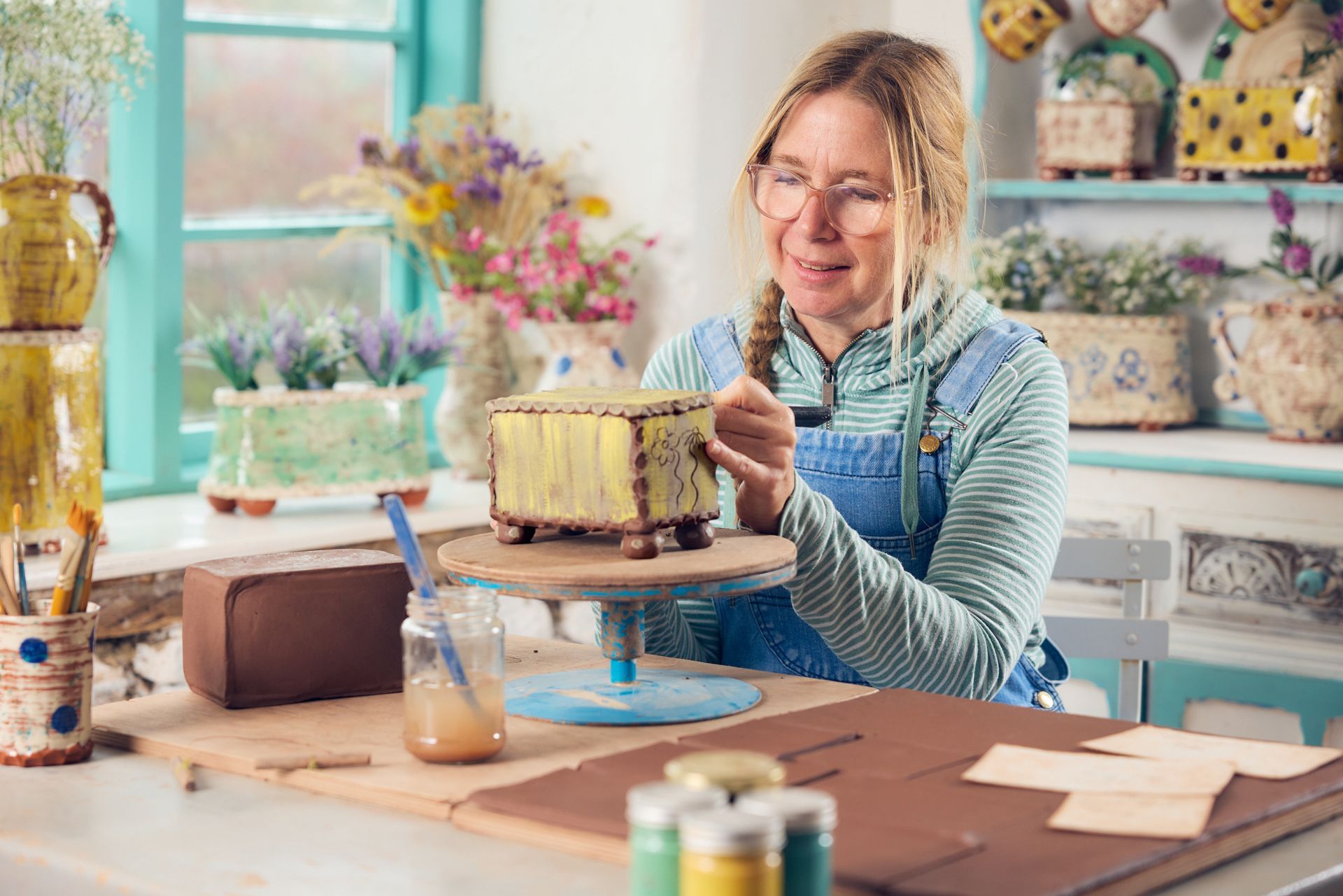 Ceramic artist Sarah Monk sat by a window, at her work table at Eastnor Pottery. Sarah has blond hair with a side plait, she is wearing pink glasses and smiling down out the pottery flower brick she is currently making. Sarah Monk is smiling slightly but is looking down, involved in the work she is doing. Sarah is surrounded by her finished pots. They are very colourful; greens, yellows, whites and blues. Many of the pots have fresh flowers in them. On the table there are 4 small jars of paint, some rolled out terracotta clay on a board, pieces of card, a pot of brushhes and a jar of water. The piece of pottery Sarah is working on is rectangular in shape and painted yellow, she is etching flowers on to the surface. The piece of pottery has been placed on a banding wheel.
