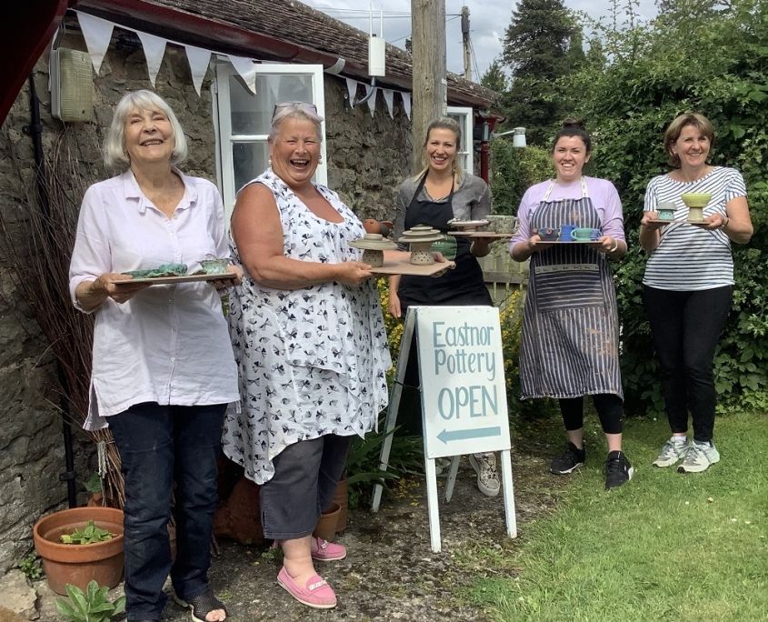 five women holding wodden boards with ceramic creations they have made standing outside eastnor pottery next to the eastnor pottery sign