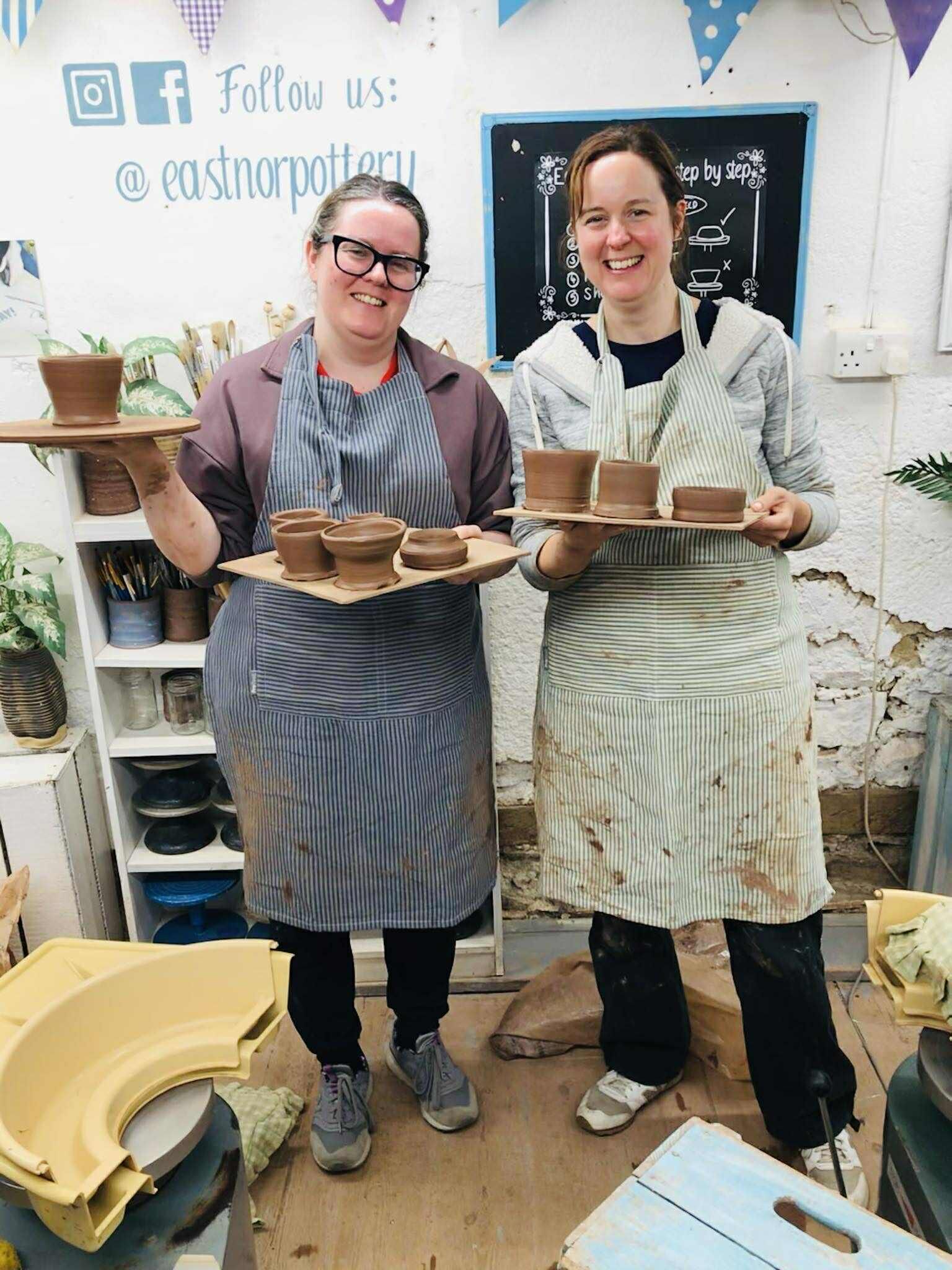 Two women standing in the pottery studio. They are wearing aprons that are covered in clay splatters. They look very happy and are holding up square boards of wet pots they have made.