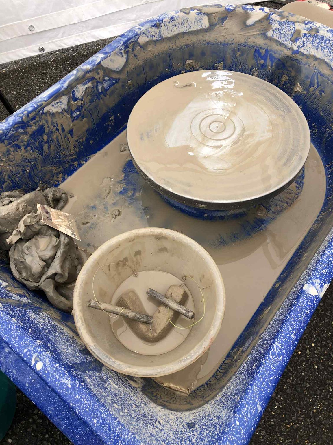 ariel view of a potter's wheel covered in clay and water. There is a small clear plastic bowl containing a cheese wire and small sponge.