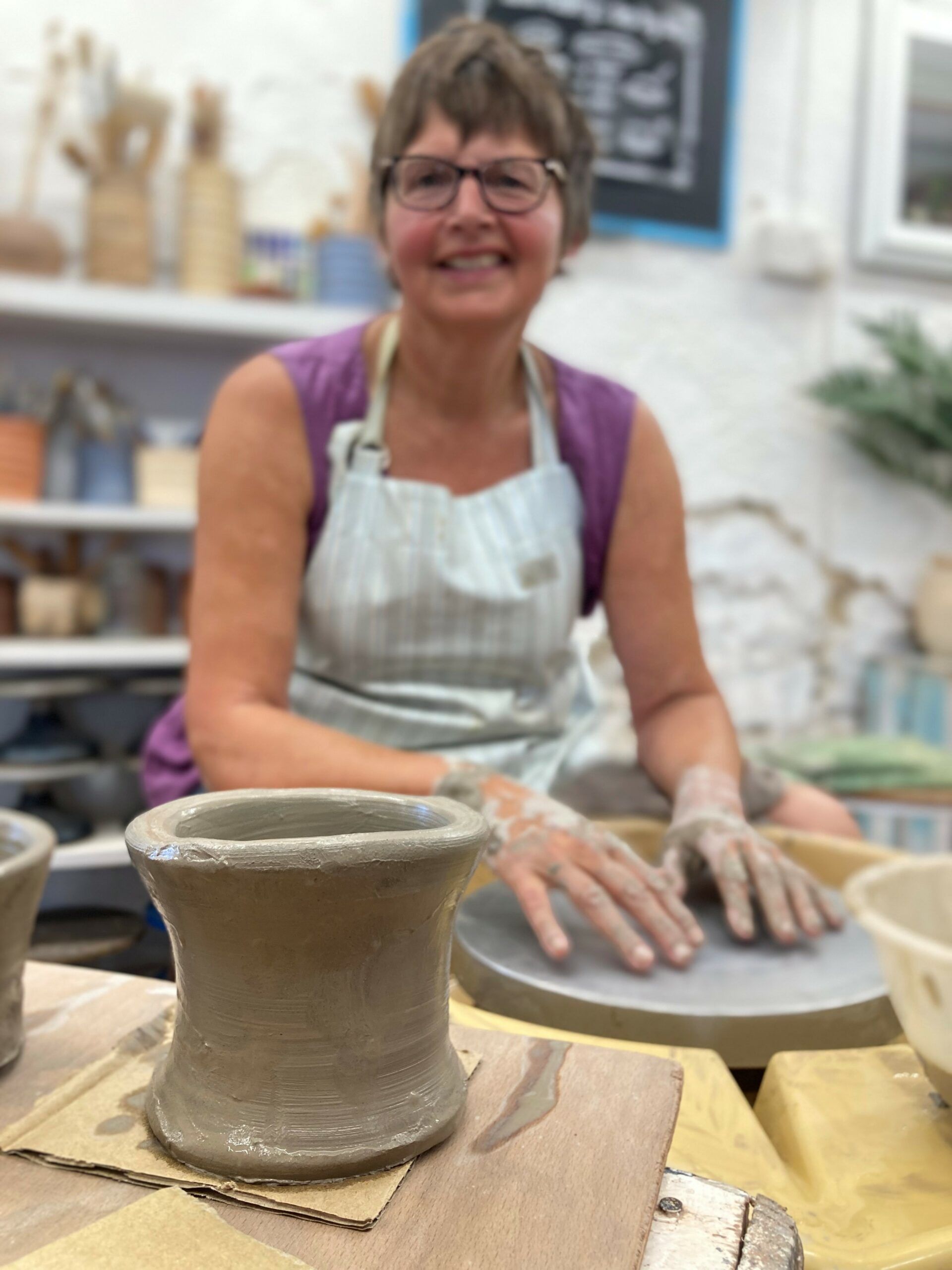 A portrait shot of class participant Gilly. She is sat at a potter's wheel in the studio at Eastnor Pottery. She is wearing a stripey apron and is smiling contentedly. Gilly's hands are resting on the wheel head and she is looking into the camera. Her hands are covered in grey clay. To the for front of the photo is Gilly's freshly thrown pot. A tall cylinder that flutes out at the top slightly.
