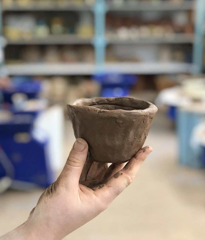 Hand shot holding a coil pot bowl up in the air, the background is of the studio shelves and pottery wheels but the background is out of focus.