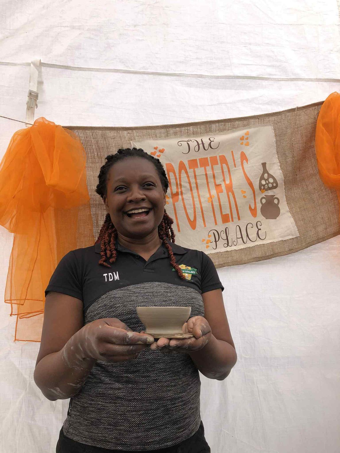 smiling woman holding a little clay bowl in both hands freshly made on the potter's wheel. There is a hand made banner fashioned from hessian and sash material in the back ground with the words The Potter's Place displayed on it