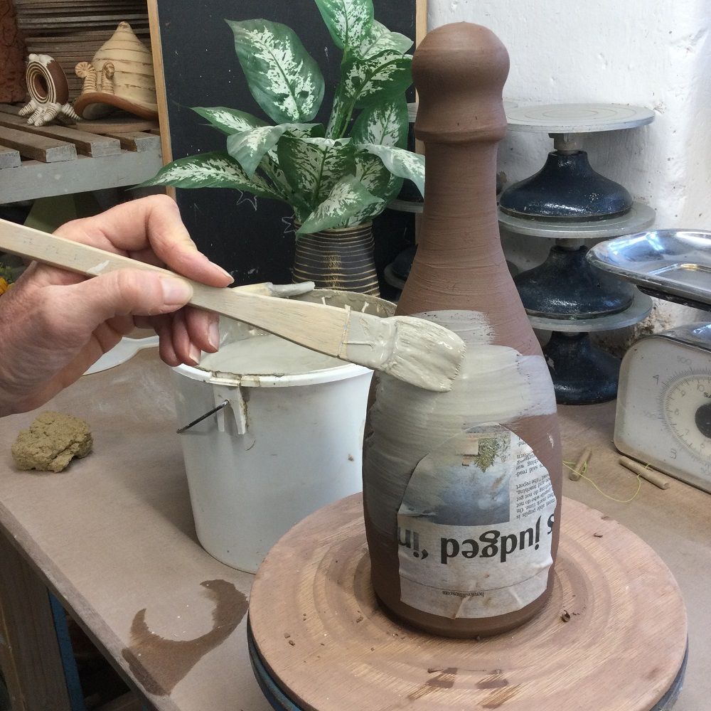 painting brush applying white slip to a hand thrown terracotta bottle