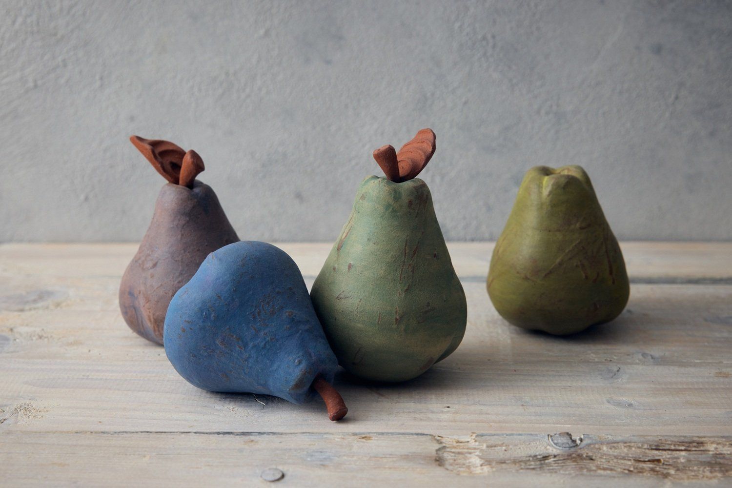 A collection of 4 pears made by Jon Williams. From left to right; a purple pear with a terracotta stalk and leaf, a blue pear lying on it's side with a terracotta stalk, a dark green pear with terracotta stalk and leaf, a light green pear with no stalk.