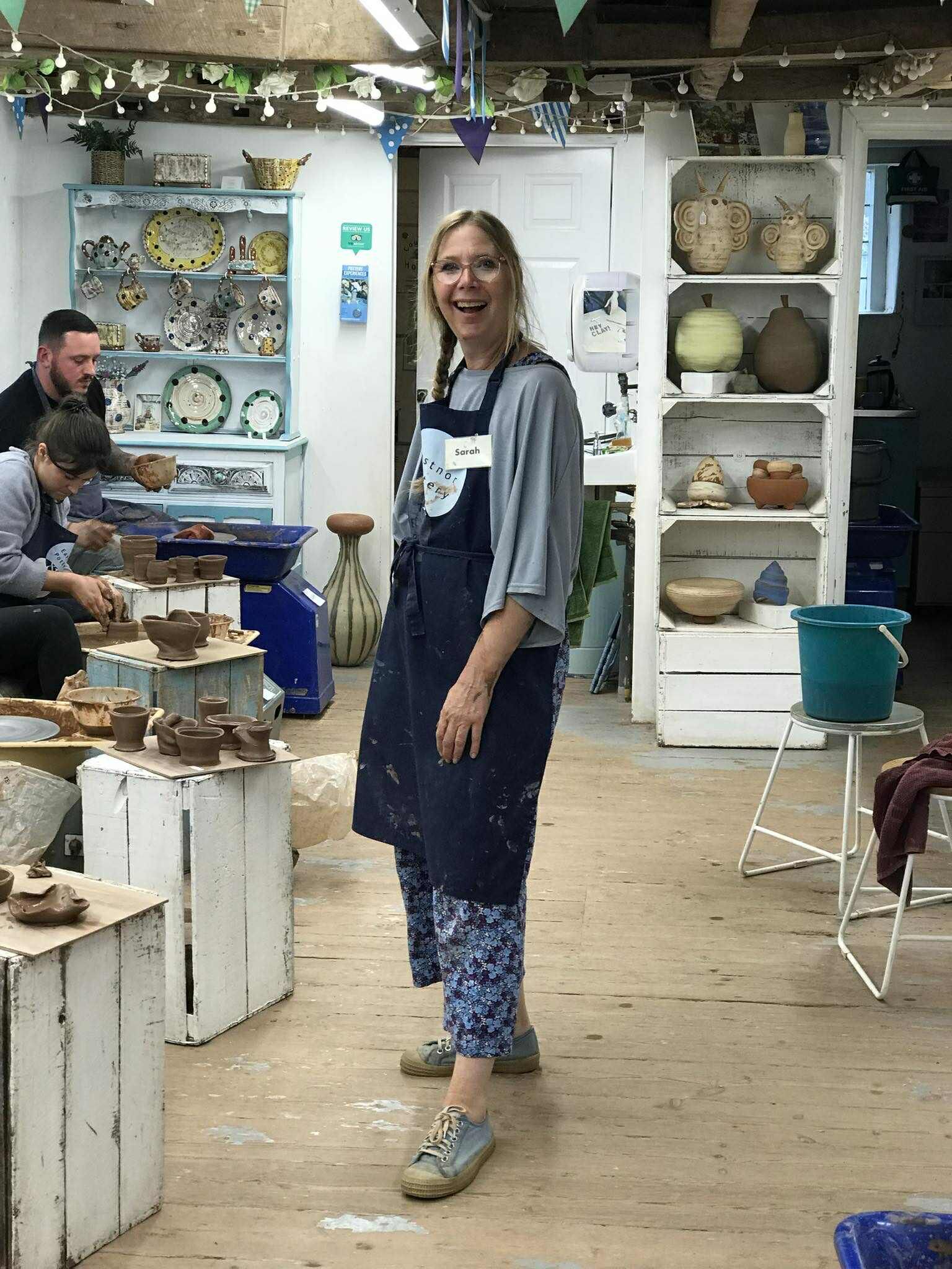 A smiling lady stood in the middle of the pottery studio surrounded by potters wheels and freshly made pots.. She is wearing an Eastnor Pottery apron with the name tag Sarah.