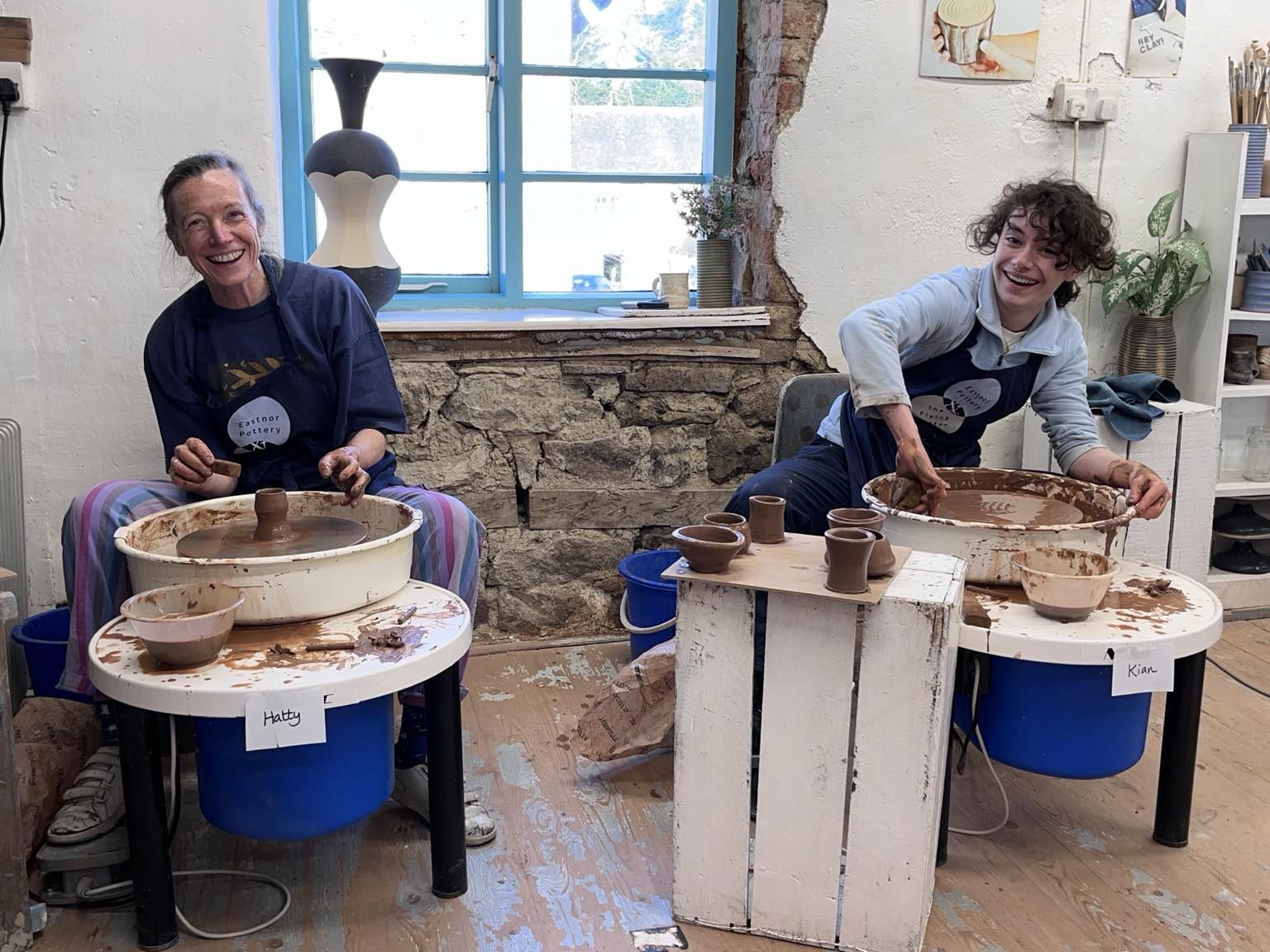 teenager and his mum sat at individual potter's wheels and laughing at the camera. they are both leaning away from each other. They are very happy to be making pots on the potter's wheel at eastnor pottery and look relaxed