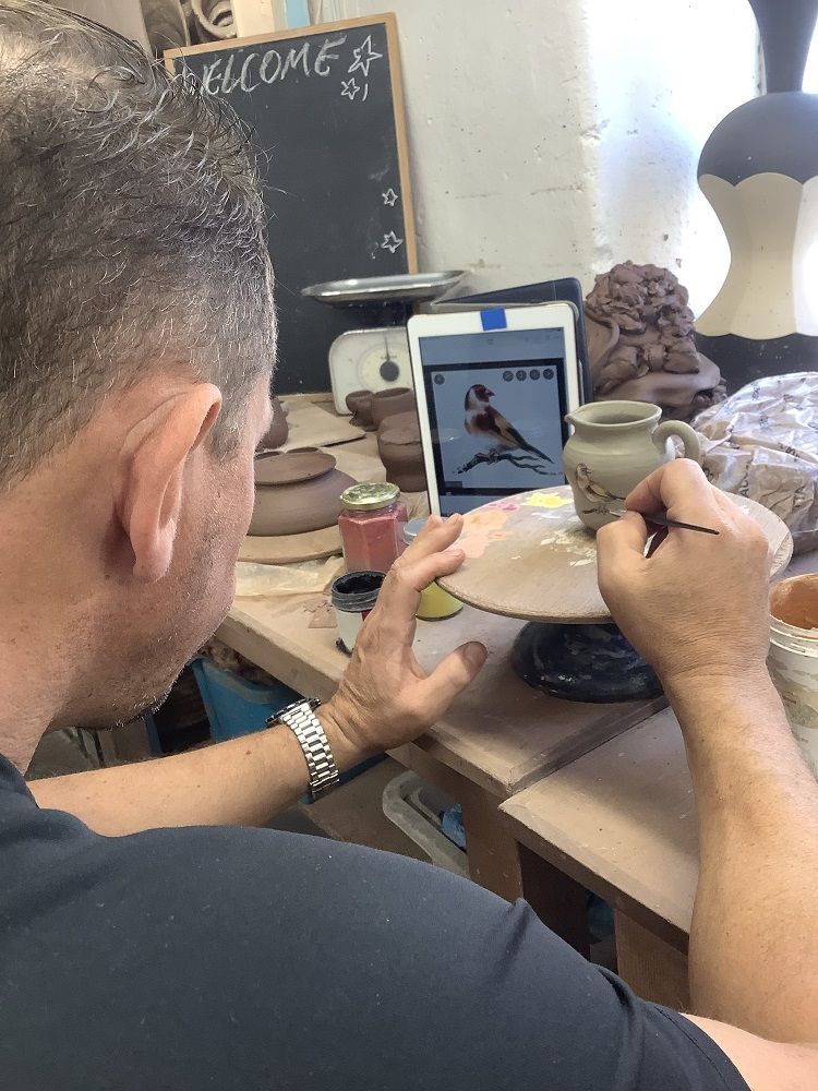 man painting a detailed image of a goldfinch bird onto the side of his handmade pot at eastnor pottery in herefordshire. very finely painted copying a photo on an ipad. The man is a course participant on a weekend potter's wheel course.