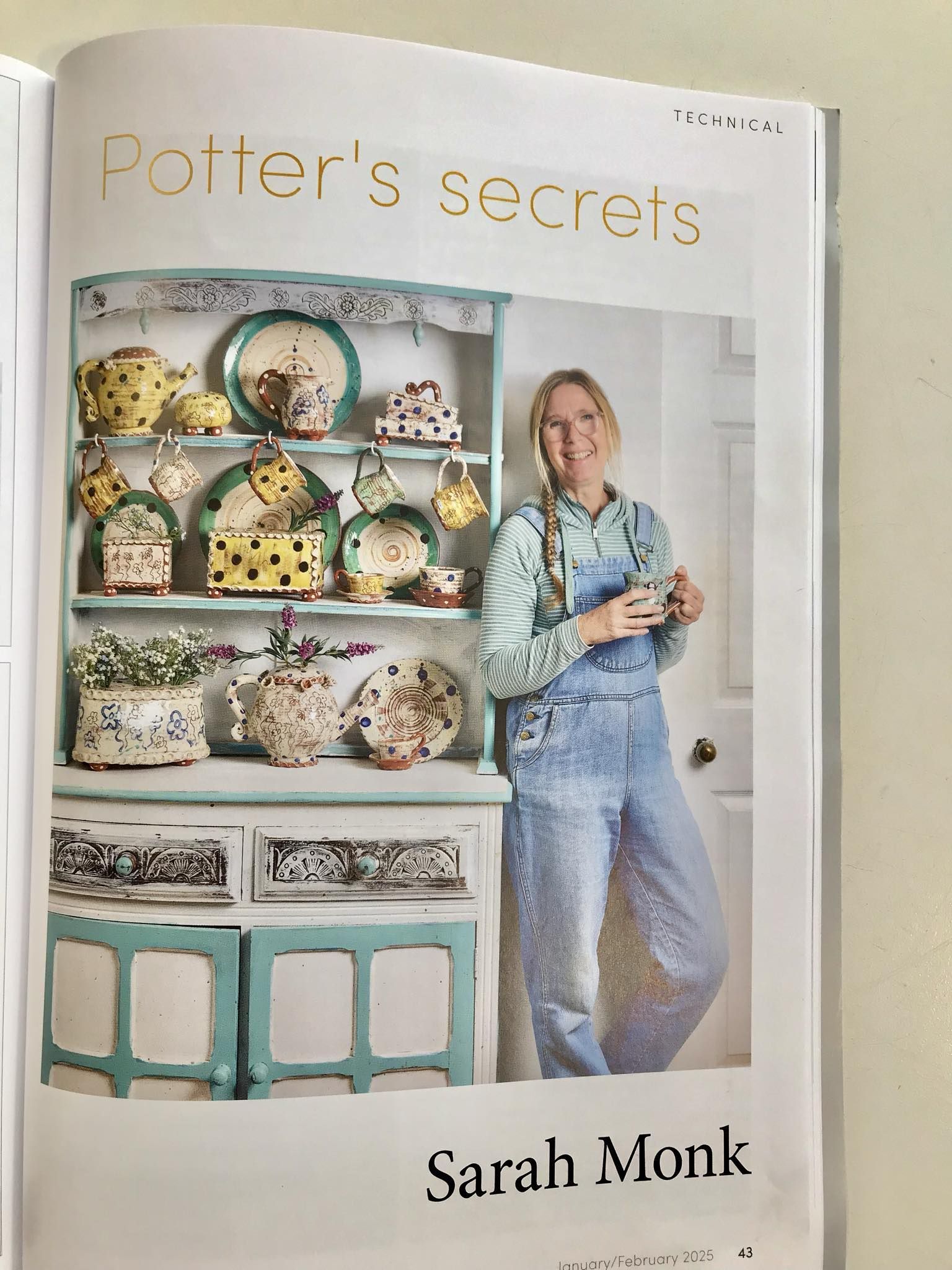 Ceramic review magazine open on page 43 showing a photograph of Sarah Monk standing in her studio at Eastnor Pottery leaning against a dresser full of her recent ceramics. Sarah is wearing blue denim dungarees, a green hoody, holding a mug and smiling at the viewer.