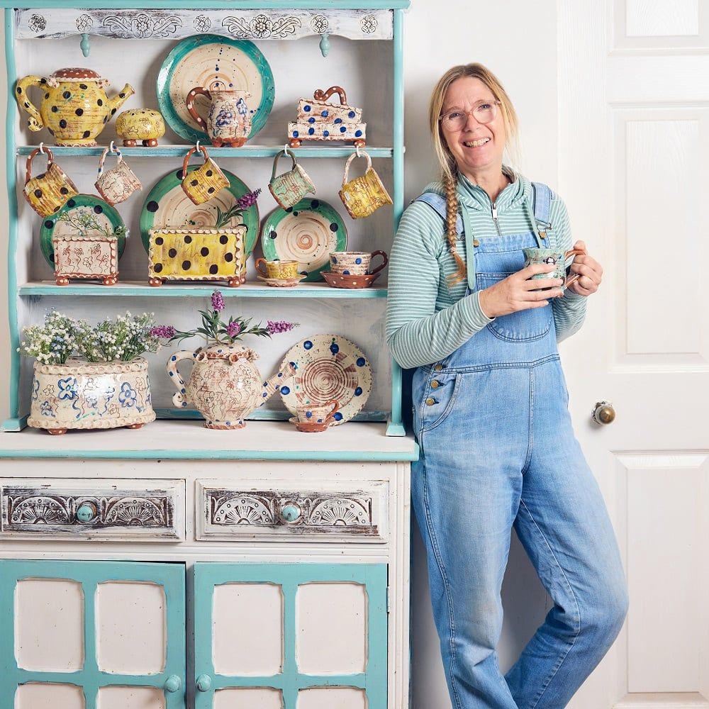Sarah Monk potter, stood in here studio at Eastnor Pottery. She is leaning against a dresser full of her colourful slipware pottery. She is holding one of her hand thrown mugs and is smiling to the camera.