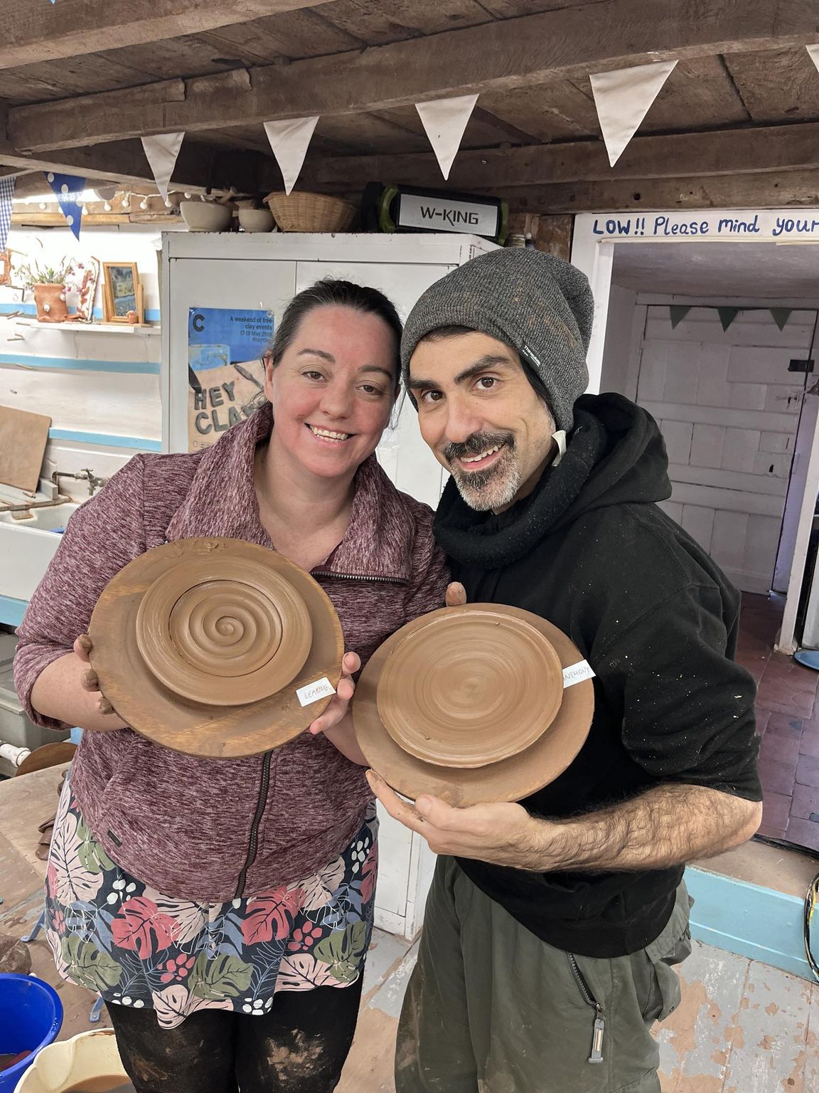 husband and wife standing close to each other smiling at the camera. Each holding a wet freshly thrown plate on a board flat to the camera. having both completed a potter's wheel course at eastnor pottery herefordshire